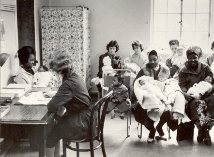 Black and white photo of manor gardens in the 1960s. Several women are sat in a waiting room holding young babies wrapped in blankets. A member of staff is writing on a file