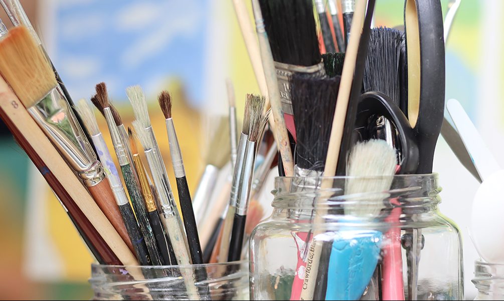 Photo of paintbrushes in a jar and colourful paintings in the background